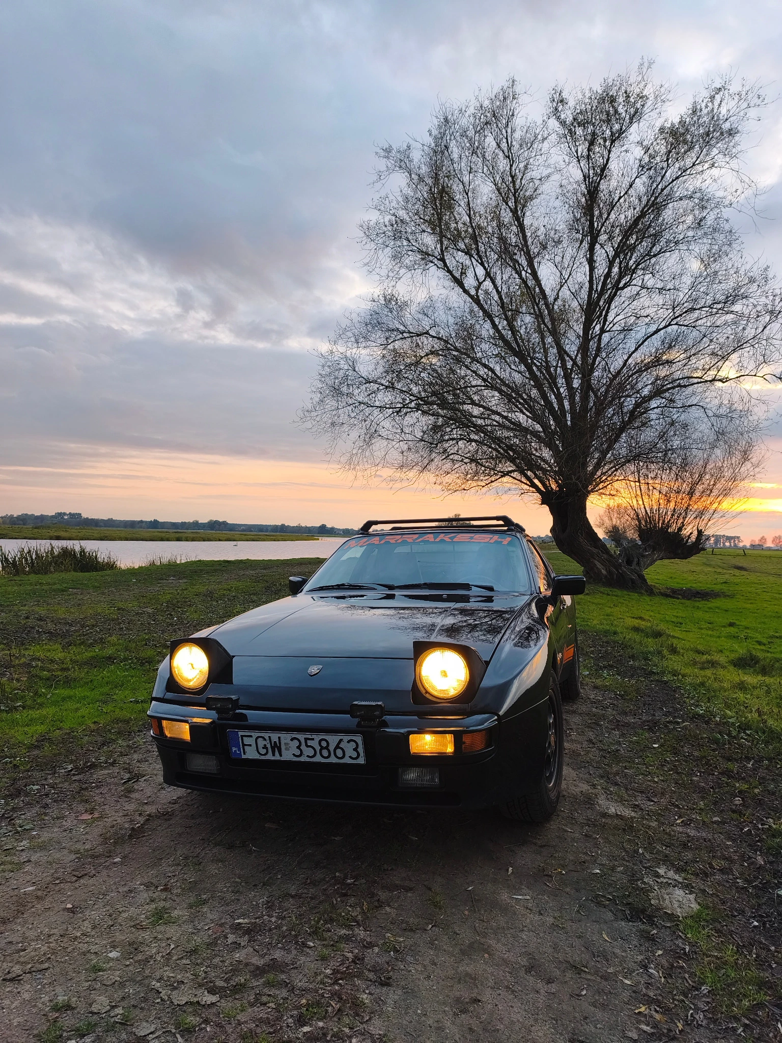 Schwarzer Porsche 944 Marrakesh mit leuchtenden Klappscheinwerfern am Feldweg im Abendlicht